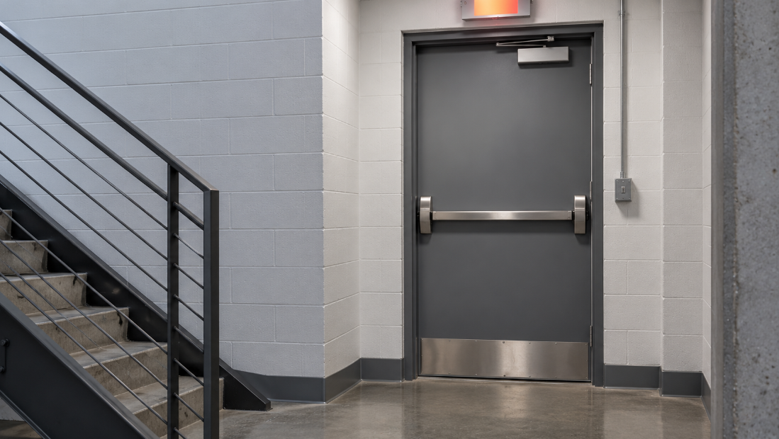Gray steel exit door with a horizontal stainless panic bar, an exit sign glowing above the frame